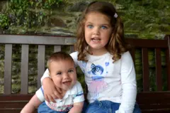 James e Kate sentados em um banco de madeira em ambiente externo. Kate tem um braço em volta de James e há uma parede de pedras com folhagem ao fundo.