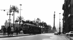 Foto em preto e branco de Barcelona em 1969, com uma rua, um ônibus passando, várias pessoas atravessando e, ao fundo, a estátua de Colombo.