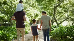 Família de quatro pessoas caminha em uma trilha na floresta, com uma criança nos ombros do pai