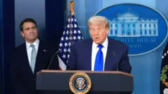 紺色のスーツに鮮やかな青色のネクタイを締めたトランプ氏が演壇に立ち、口を開いている。後方には、ダークスーツに水色のネクタイを締めたブランチ氏がいる