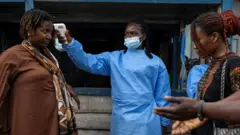 Em uma rua na cidade de Goma, na República Democrática do Congo, um profissional de saúde com máscara facial azul e avental médico mede a temperatura de uma mulher com um dispositivo próximo à cabeça. Outra mulher e um homem estão do outro lado esperando.