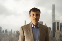 Sharma usando um terno marrom e uma camisa azul clara em baixo. Atrás dele há vários prédios