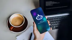 صورة من أعلى ليد سيدة تجلس أمام لابتوب، وأمامها كوب من القهوة، وتمسك بهاتف ذكي يظهر عليه صورة روبوت دردشة كتبت تحتها: Hello! Ask me anything.. 