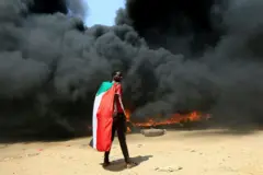 صورة أرشيفية لشخص يرتدي علم السودان يقف أمام كومة من الإطارات المشتعلة خلال احتجاج ضد احتمال الحكم العسكري في الخرطوم، السودان، 21 أكتوبر/تشرين الأول 2021.