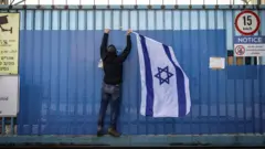 إسرائيلي يعلق علم إسرائيل على البوابة المغلقة لمبنى وكالة الأونروا في القدس الشرقية المحتلة في 30 يناير/كانون الثاني 2025