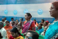 Mujer de pie sosteniendo un micrófono en medio de una reunión comunitaria, rodeada de varias personas sentadas en sillas de plástico, con un fondo azul y blanco que muestra logotipos circulares.