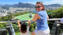 Rebecca sorrindo em paisagem panorâmica do Rio; sua filha está de costas