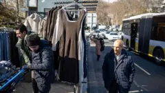 در یک خیابان دستفروشی پوشاک برای فروش گذاشته و چند عابر در حال عبور و مرور هستند