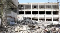 ساختمانی سفید با پنجره‌هایی شکسته پس از حمله اسرائیل به زندان اوین در ایران دیده می‌شود. توده‌ای از آوار، شامل قطعات فلزی و بتنی، در مقابل ساختمان انباشته شده است. در پیش‌زمینه، شاخه درختی به چشم می‌خورد.