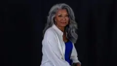 Shirley Chung usa uma camisa branca em cima da uma blusa azul. Ela tem cabelos longos e grisalhos