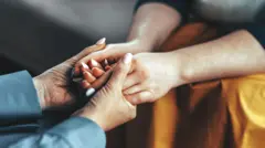 As mãos de uma mulher seguram as de outra que está sentada à sua frente