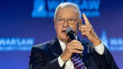 Кіт Келлог тримає мікрофон та підняв вказівний палець вгору