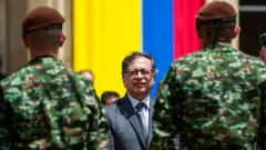 O presidente da Colômbia, Gustavo Petro, de terno, com a bandeira do país ao fundo e dois soldados à frente