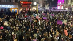 Kalabalık bir kadın topluluğu akşam karanlığında ellerinde dövizlerle toplanmış, arkada polisler zırhlarıyla sıralanmış