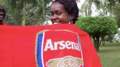 Arsenal fan Agnes Katende wrapped in an Arsenal flag looks over her shoulder at the camera in a garden in Kampala.