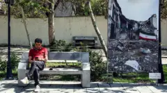 یک مرد ایرانی هنگام نشستن روی نیمکتی و نگاه کردن به تلفن همراهش، در کنار یک عکس بزرگ که «جنگ کنونی خاورمیانه» را نشان می‌دهد، در پارکی در تهران دیده می‌شود؛ این تصویر در ۱۶ آوریل ۲۰۲۶ (۲۷ فروردین ۱۴۰۵) ثبت شده است. قطع اینترنت که اندکی پس از حمله آمریکا و اسرائیل به ایران در ۲۸ فوریه ۲۰۲۶ (۹ اسفند ۱۴۰۴) اعمال شد، طبق گزارش نت‌بلاکس تا ۵ آوریل (۱۶ فروردین ۱۴۰۵) طولانی‌ترین قطعی سراسری ثبت‌شده در کشور بوده و اکنون بیش از ۵۰ روز ادامه یافته است. اینترانت بسیار محدود داخلی ایران در طول جنگ فعال بوده و به مردم امکان اتصال به وب‌سایت‌های داخلی را می‌دهد، اما همچنان برای فعالیت عادی کسب‌وکارها در کشوری که اقتصاد آن پیش‌تر نیز تحت تأثیر تحریم‌های بین‌المللی تضعیف شده، کافی نیست.