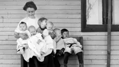 Black and white photo showing a woman from around the turn of the century on a bench, with four babies in her arms. Another baby is wedged in between her and a toddler, who is hanging his legs off the bench. She wears a white, high-necked blouse and long black skirt, while her hair is in a high bun.