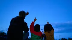 Três pessoas e um homem sentados ao ar livre olhando e apontando para o céu