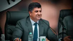 لاهور شيخ جنكي يجلس على مكتب وأمامه مكيروفون، مرتدياً سترة رمادية اللون وربطة عنق زرقاء دوائر صغيرة بيضاء، وقميصاً أبيض.