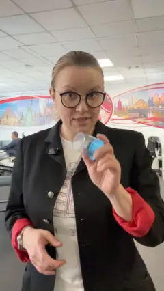 Novinarka drži čašicu za testiranje urina u ruci i objašnjava šta boja urina govori o zdravlju.