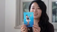 Arden Cho sorri para a câmera, segurando uma fotografia de si própria quando criança. Ela tem longo cabelo castanho-escuro ondulado e, na fotografia, está em uma apresentação de dança, vestindo uma fantasia com saia e mangas bufantes, em pose com as pernas cruzadas e as mãos no ar.