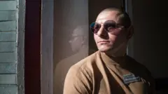 Andrii Askerov, de camisa marrom e óculos escuros, olha pela janela de uma prisão.