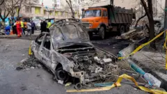 Yanmış ve harabeye dönmüş bir kar yol kenarında park etmiş. Geri planda bir grup insan toplanmış. 