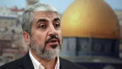 Khaled Meshaal stands in front of an image showing the Dome of the Rock while wearing a white shirt and black blazer.