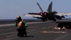 A man kneels down on the deck of aircraft carrier USS Gerald R Ford as a fighter plane is seen behind him (09/03/26)