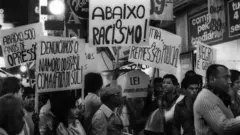 Foto preta e branca que mostram várias pessoas em uma manifestação com faixas "Abaixo o racismo"