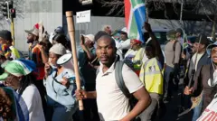 man hold stick while protesting, South Africa flag dey show for di side