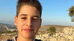 محمد إبراهيم مراهق فلسطيني يحمل الجنسية الأمريكية يبتسم في صورة شخصية له، ويصفف شعره الكستنائي إلى الأعلى. خلفه في صورة التقطت نهاراً أرض مليئة بالحجارة وعدد من الأشجار الصغيرة.