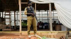 Um policial armado, vestindo uniforme cáqui e colete de proteção contra perfurações preto, faz a guarda em frente a uma instalação improvisada