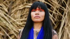 Fotografia colorida mostra uma mulher do povo Yawalapiti, com cabelos lisos e negros, usando uma pintura ritual vermelha e colar de miçangas azuis