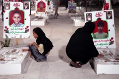 Una mujer y una niña con velos negros agachadas junto a las tumbas de los niños muertos en el ataque a la escuela de Minab. Detrás de las tumbas se exhiben imágenes de los rostros de los niños, sobre fondos rojos.