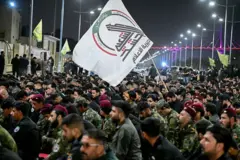 Membros das Forças de Mobilização Popular (FMP) reuniram-se em Basra, no sul do Iraque, para uma cerimônia em memória do líder iraniano, o aiatolá Ali Khamenei, morto em ataques aéreos dos EUA e de Israel. Na fotografia, eles carregam faixas com a imagem de Khamenei, bandeiras das FMP e do Kataib Hezbollah, e entoam slogans em apoio ao Irã e aos grupos de resistência islâmica.