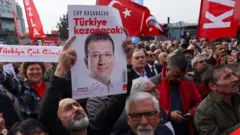 Много людей с разными плакатами, в центре мужчина держит белый плакат с потретом Экрема Имомоглу и надписью на турецком языке.