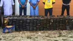 Ifoto iterekana mu maso igaragaza abagabo batanu bahagaze, imbere yabo hari ibirundo bya cocaine