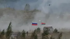 白煙の立ち上る野原に木々と小屋が点在し、両国国旗の隣に兵士が1人立っている。約10メートル上にはドローンが1機飛んでいる