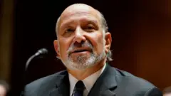 濃い灰色のスーツを着たラトニック長官が前を向いて話している