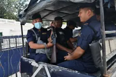 नेपाल प्रहरीको टोली काठमाण्डूको सिंहदरबार परिसर छेउमा प्रहरी भ्यानमा ड्यूटीमा। पछाडिको सीटमा बसेका एक बर्दीधारी प्रहरीले मुखमा नीलो मास्क लगाएका छन् र हातमा बन्दुक बोकेका छन् 