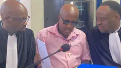 Umugabo yambaye ishati y'iroza (ibara ry'umwambaro w'abanyororo) hagati y'abunganizi 2 bari mu mwambaro w'abashingwamanza