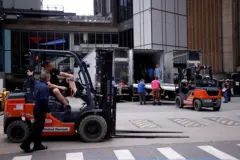 Do lado de fora do Madison Square Garden, trabalhadores descarregam diversos caminhões estacionados em frente à arena. É possível ver equipamentos sendo transportados por empilhadeiras