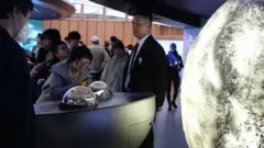 Visitors look at moon samples encased on a table at an expo. There is a large sculpture of the moon near them.