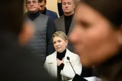 Юлія Тимошенко у залі суду сидить у білому пальті та слухає, дивлячись вперед. Навколо неї та на першому плані - фігури інших людей