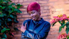 Umuririmvyikazi Marlène Kamikazi batazira “B Charly” yambaye ikote ry'umukara, inyuma ye hari inzu y'amatafari, iruhande rwe hari amashugwe. 