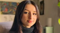 Aisha Belarbi looking directly down the camera. She is wearing a black leather jacket with a dark grey turtle neck sweater underneath. She is smiling, though her teeth are not showing. Her straight dark brown hair is parted in the middle of her head and is worn down, falling below her shoulders. She is lit from the right of the image. She is indoors, though the background of the picture is blurred.