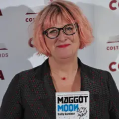 Sally segurando um de seus livros. Ela é uma mulher branca, usando um óculos com armação preta e cabelo rosa.