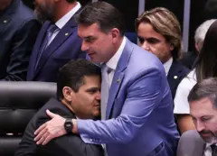Davi Alcolumbre e Flávio Bolsonaro usando ternos, em sessão do Senado, com Davi sentado em poltrona e sendo abraçado por Flávio Bolsonaro