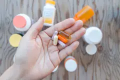 Uma mão segurando um monte de suplementos em uma palma aberta. Há frascos de suplementos como pano de fundo.